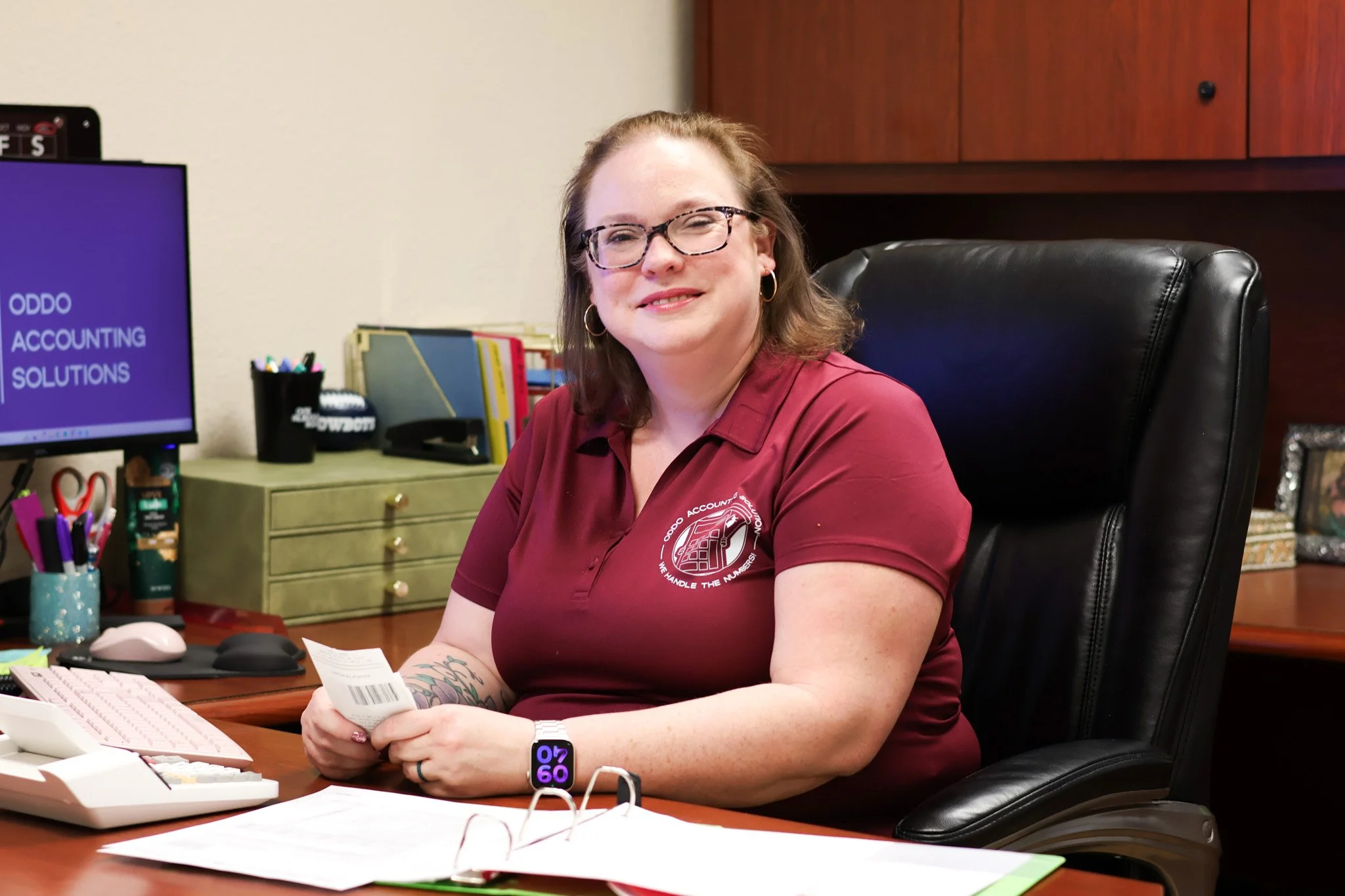 Woman with glasses and tattoos sitting at desk in office, holding receipt, smiling.