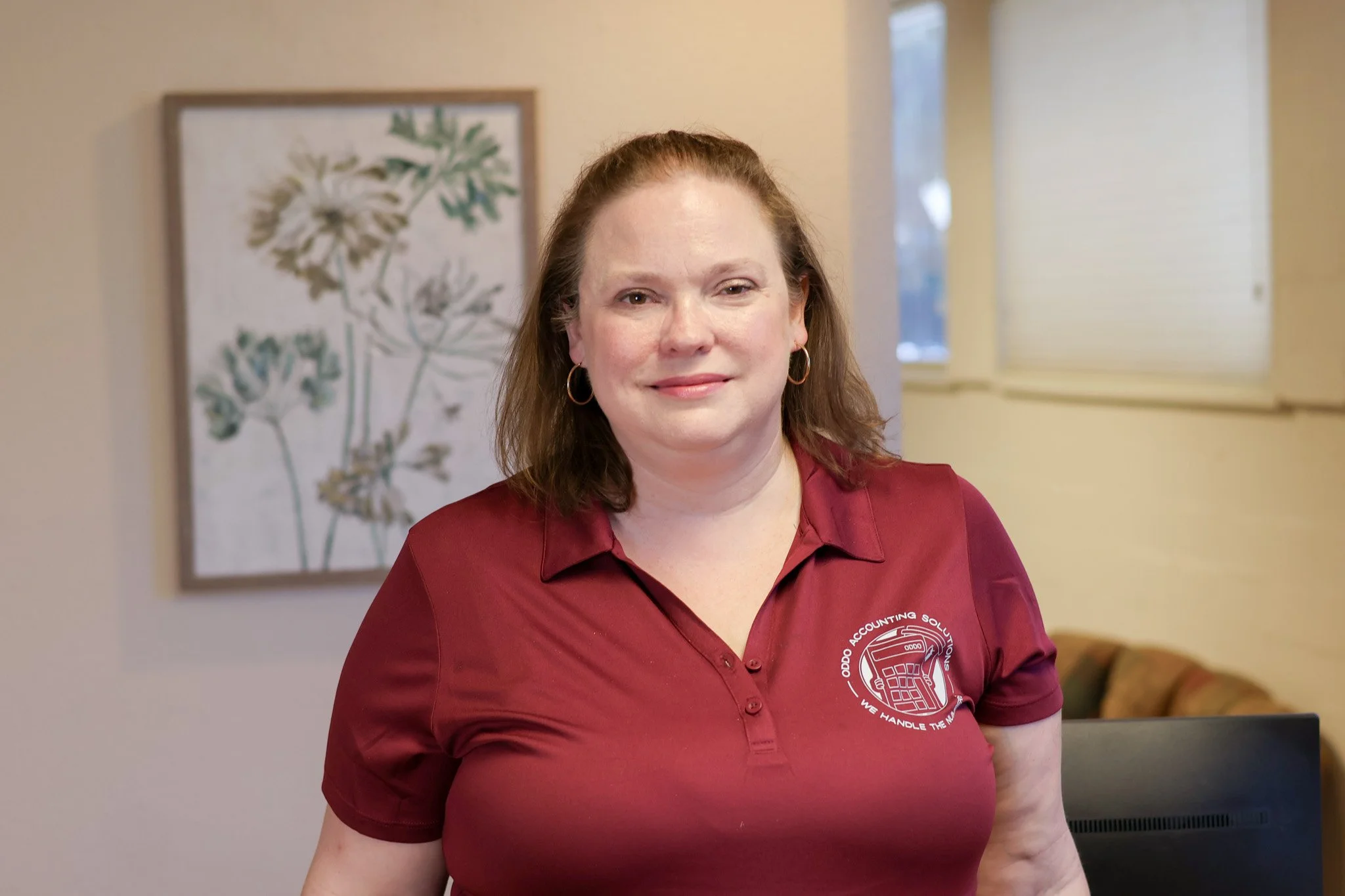 A woman with red hair wearing a maroon polo shirt with a logo, standing in an indoor setting with framed botanical artwork on the wall behind her.