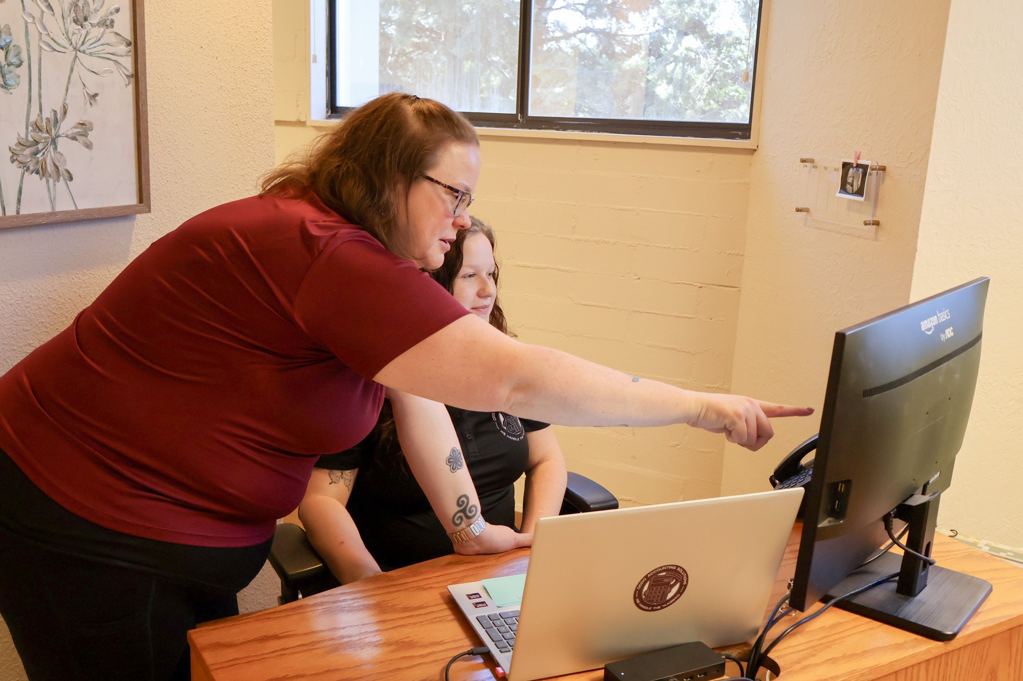 A woman with glasses and a tattoo on her arm points at a computer monitor while sitting beside a girl with tattoos on her arm. They are at a wooden desk with two laptops in a room with yellow walls, a window, and artwork on the wall.