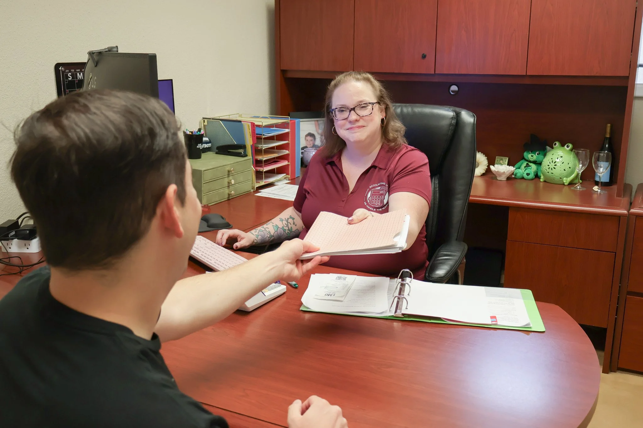 A woman with glasses and tattoos on her arm sitting at a desk, handing a folder to a man with short brown hair. The desk has paperwork, a calculator, and a binder. Behind her are decorations, including frog figurines and a wine bottle.