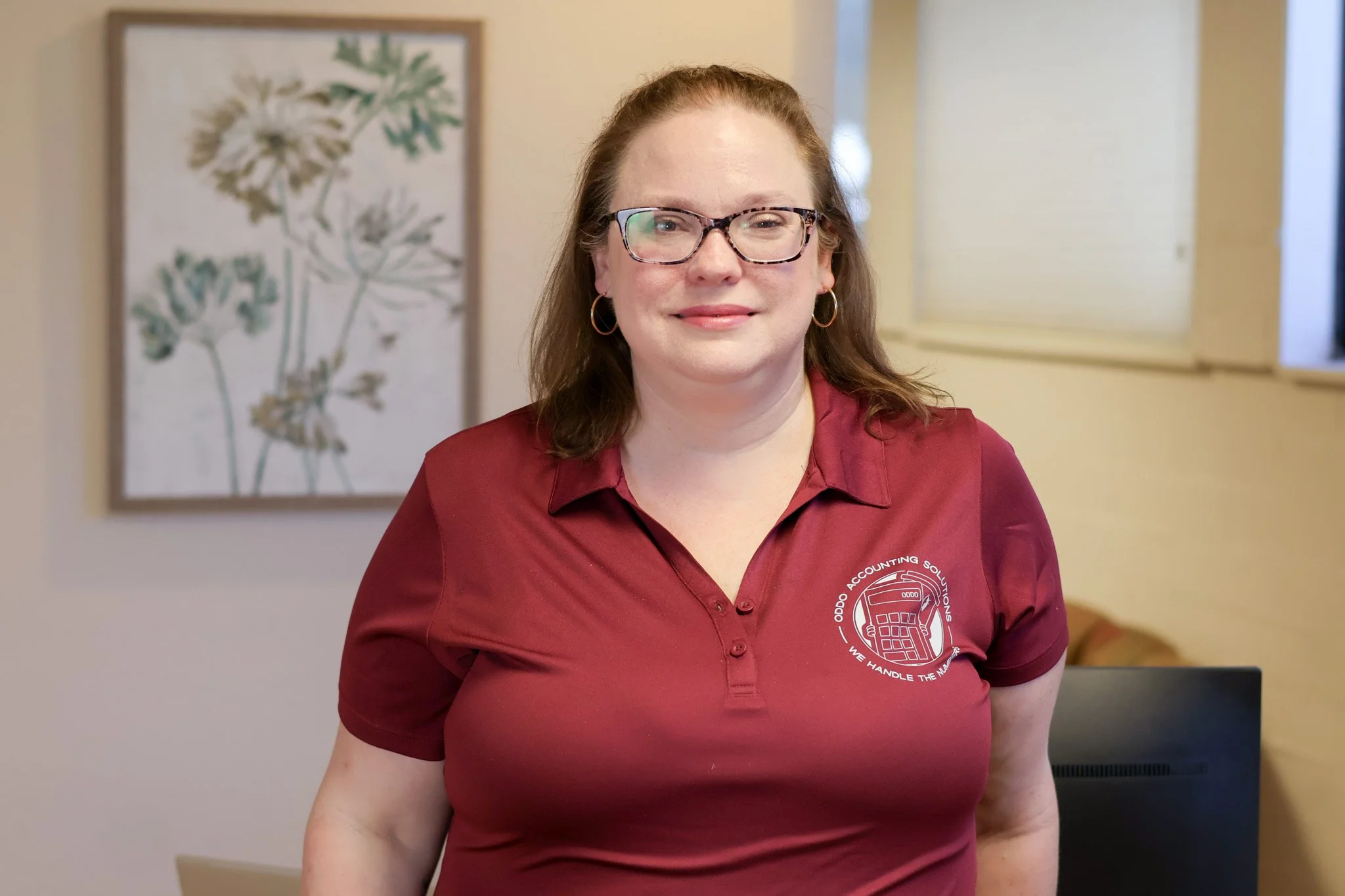 A woman with long brown hair, glasses, and hoop earrings, wearing a maroon collared shirt with a logo on the pocket. She is smiling and standing indoors with a framed botanical print behind her, illuminated by natural light.