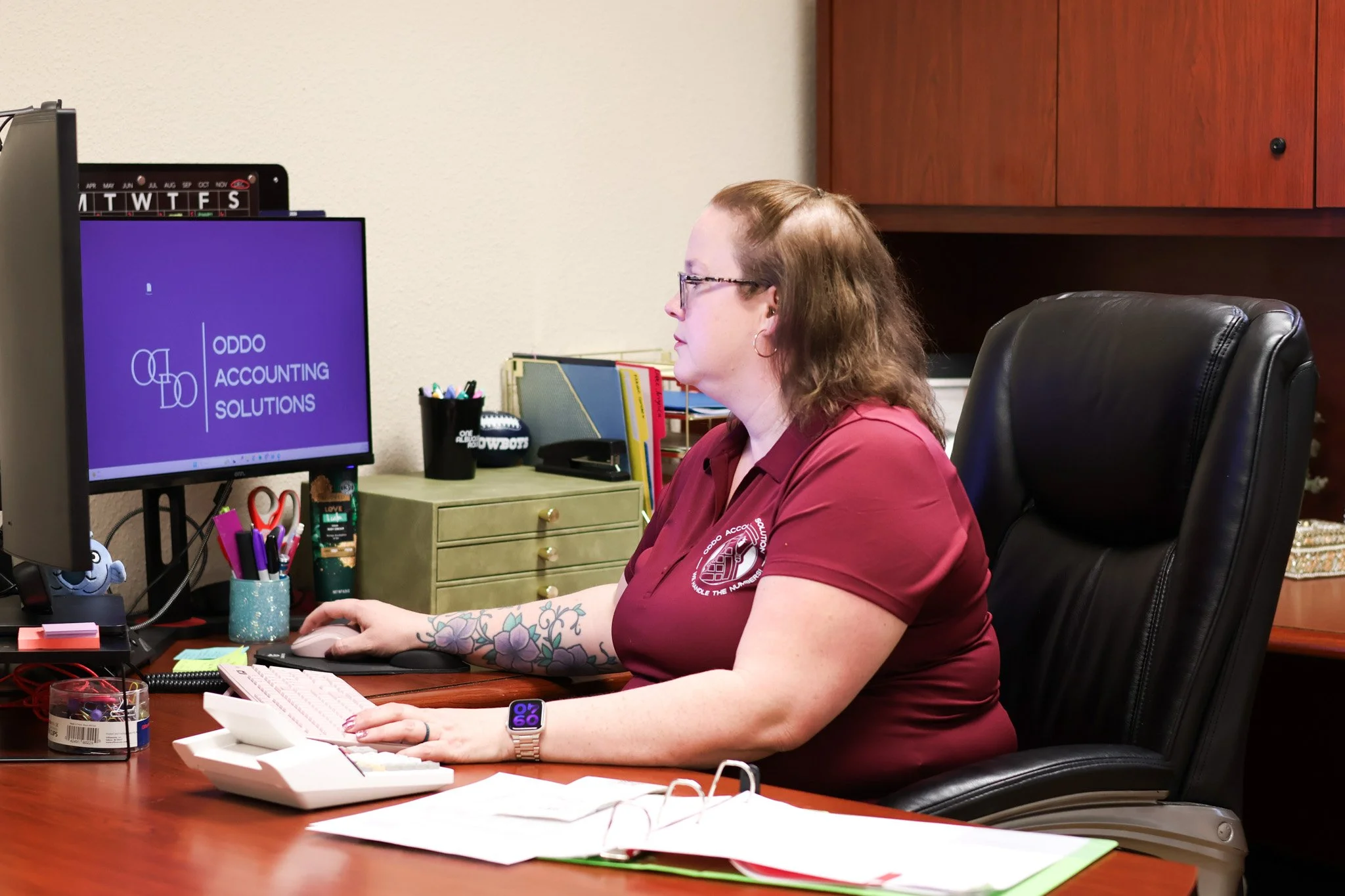 A woman with glasses and tattoos on her arm working at a desk with office supplies, a computer monitor displaying 'ODDO ACCOUNTING SOLUTIONS,' and various office items in an office environment.