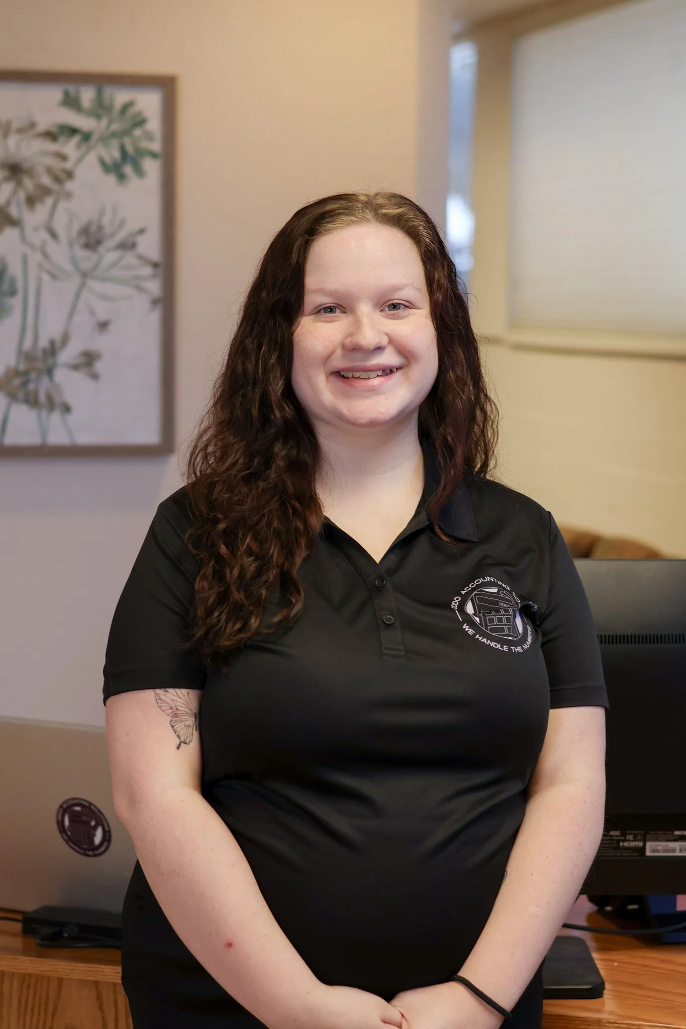 A young woman with long, curly brown hair and light skin, smiling, wearing a black polo shirt with a logo, standing indoors with a painting of green leaves on the wall behind her, and a window with closed blinds to her side.