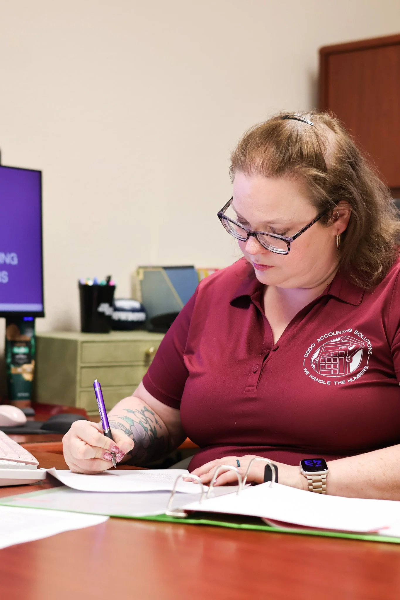 A woman with glasses and a tattoo on her arm is sitting at a desk, writing on paper. She is wearing a maroon polo shirt with a logo that reads 'ODDO Accounting Solutions' and 'We Handle the Numbers.' She has a smartwatch on her left wrist and earrings, with a pen in her right hand. In the background, there is a computer monitor, office supplies, and filing cabinets.