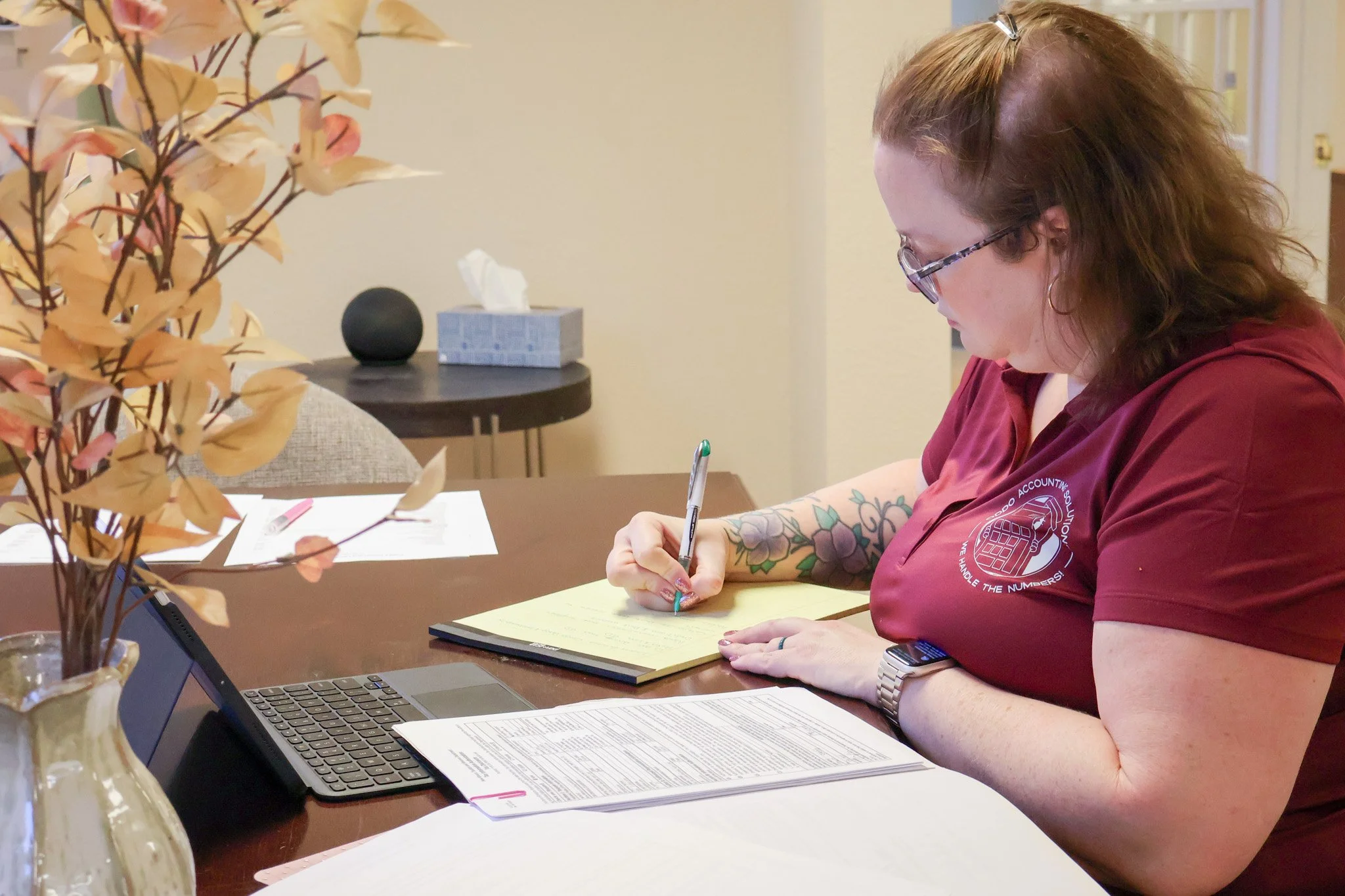 A woman with tattoos wearing glasses and a maroon shirt, sitting at a table, writing on a document with a laptop, in an indoor setting.