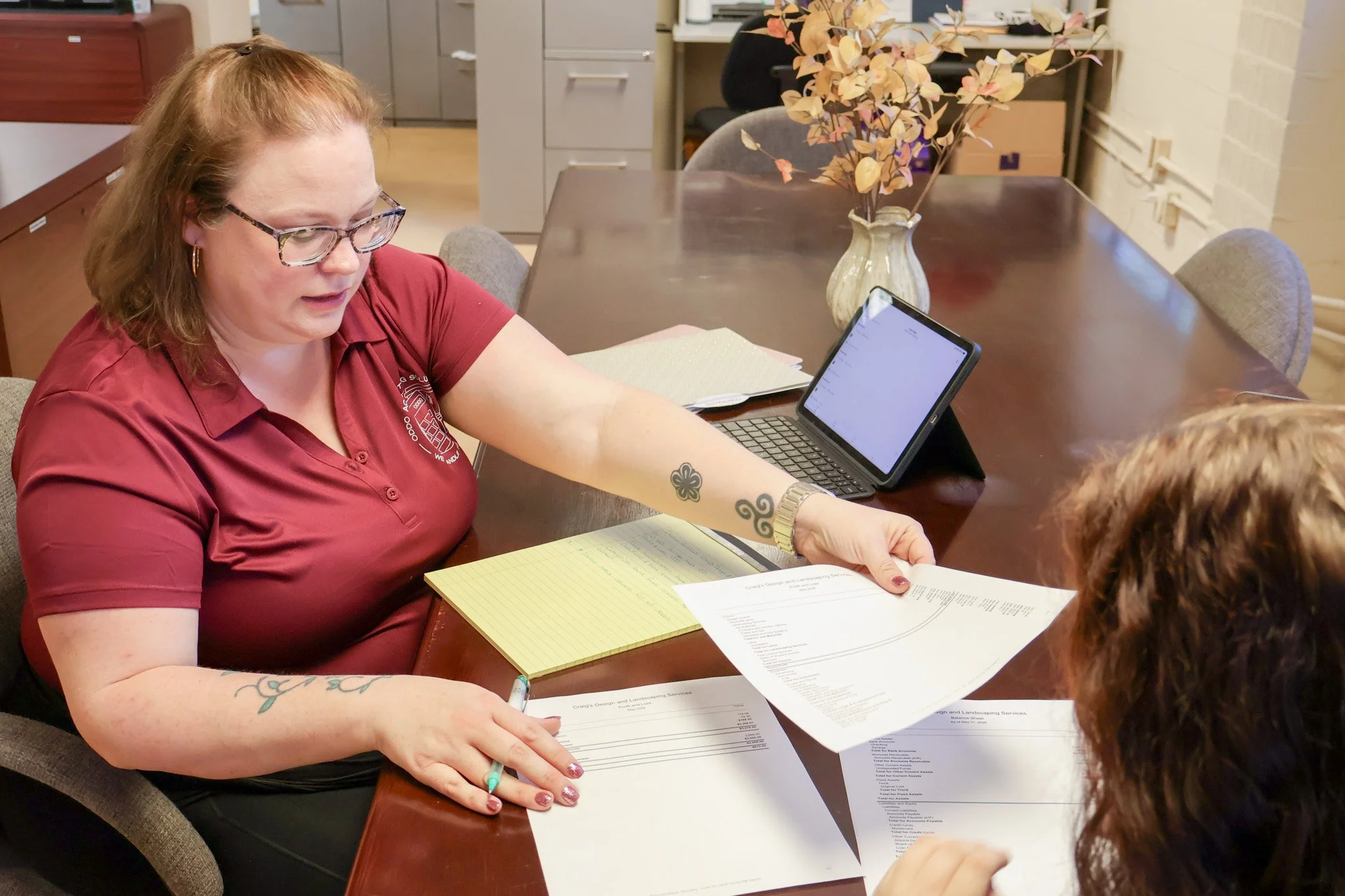 Woman wearing glasses and a maroon shirt sitting at a table, handing papers to another person with curly hair, while various documents and a notepad are on the table, with a vase of dried flowers in the background.