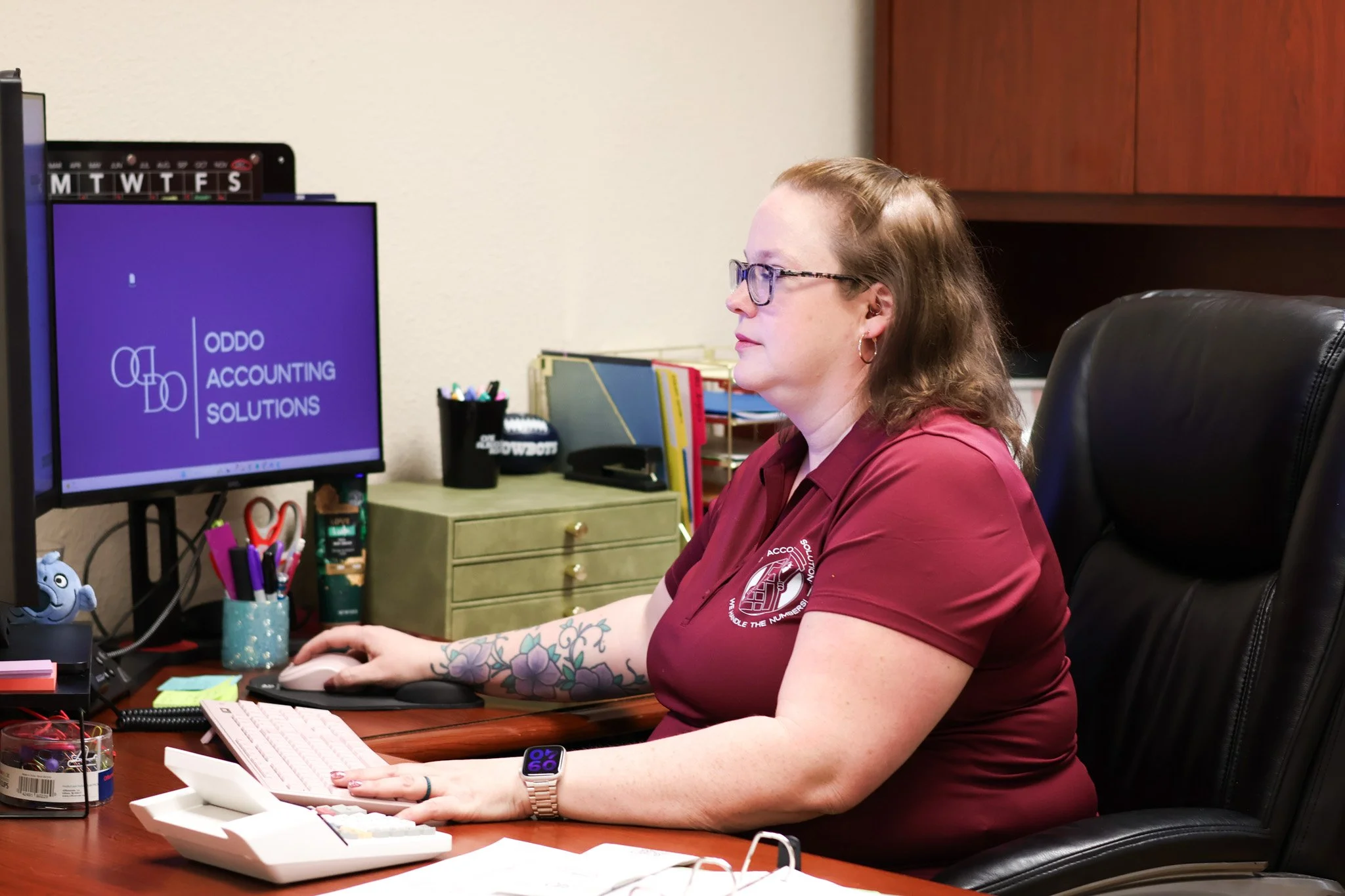 A woman with light skin, glasses, and tattoos on her arms, sitting at a desk in an office. She is wearing a maroon shirt with a logo, using a computer mouse and keyboard, with a monitor displaying 'ODDO ACCOUNTING SOLUTIONS.'