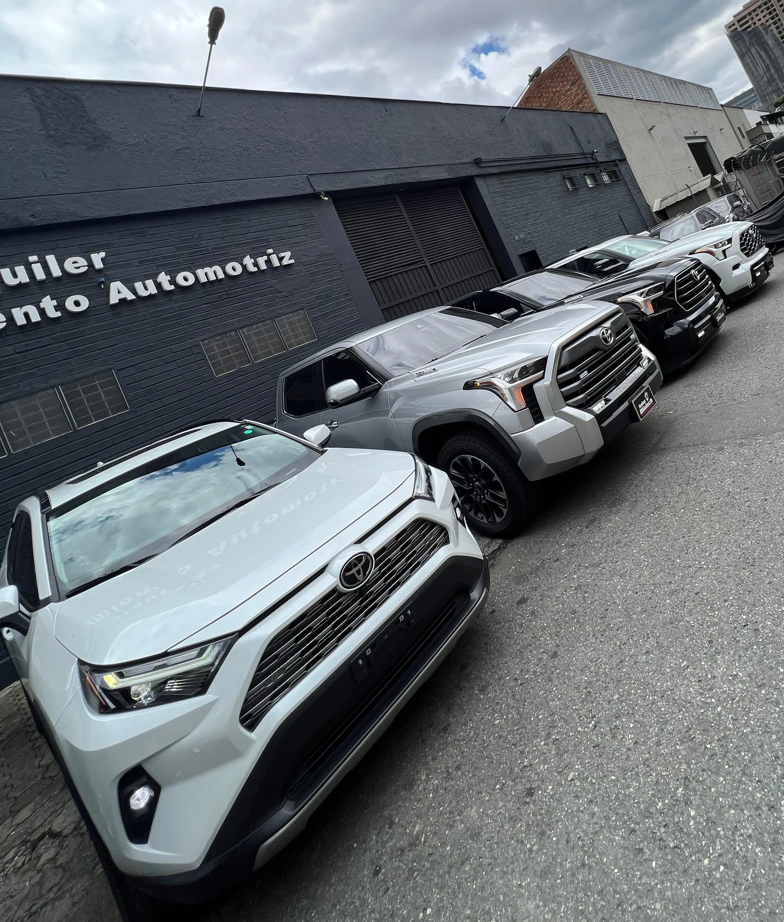 Estacionamiento con varios autos, incluyendo modelos Toyota y Toyota Tacoma, frente a un taller automotriz con un muro negro y letras blancas en la pared que dicen 'Alquiler' y 'Automotriz'.