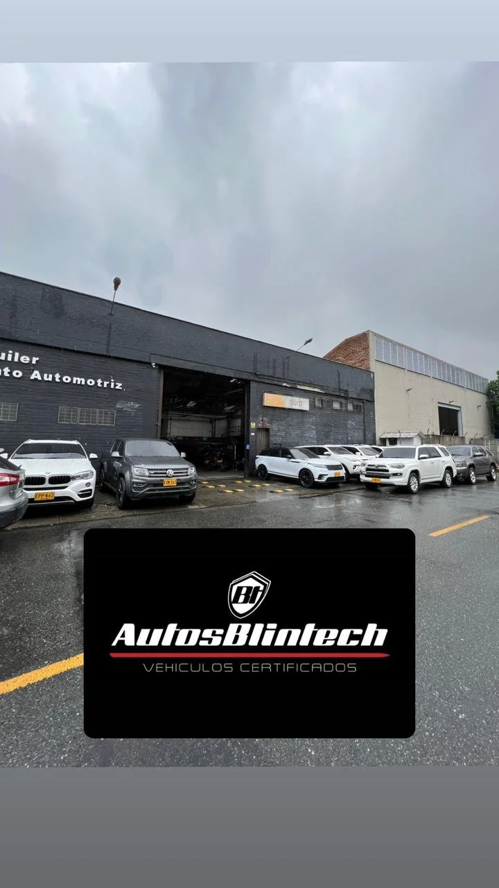 Lote de autos certificados en un taller automotriz llamado Autos Blintech, con varios autos blancos y grises estacionados en frente del taller bajo un cielo nublado.