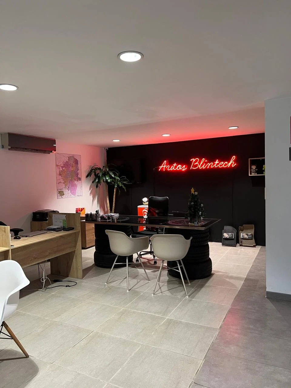 Recepción de oficina con escritorio, sillas blancas y una planta grande, con cartel de neón que dice 'Autos Blintech' y decoraciones navideñas.