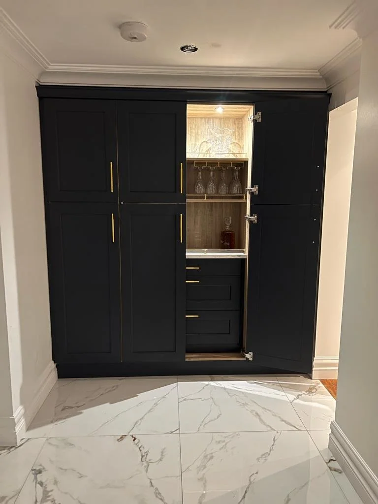 A built-in black cabinet with gold handles, with a section open revealing glassware and shelves inside, situated in a room with white marble tile flooring.