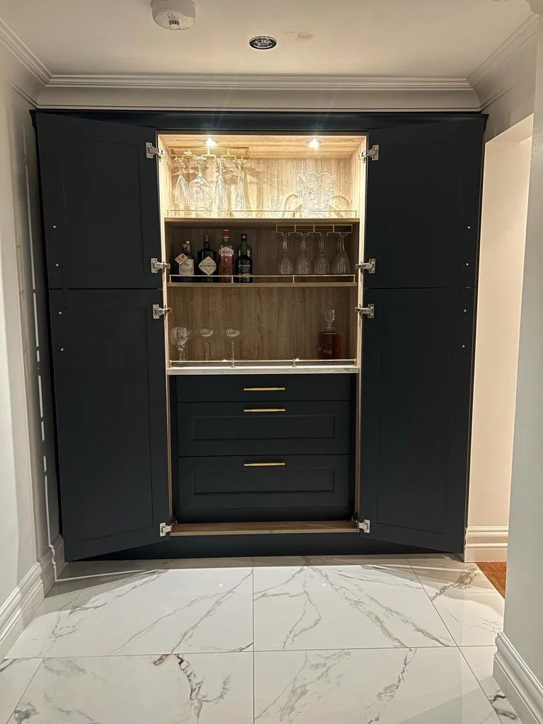 A dark blue cabinet with open doors, illuminated interior, containing glassware and liquor bottles, with drawers below, positioned against a white tiled floor with gray veining.