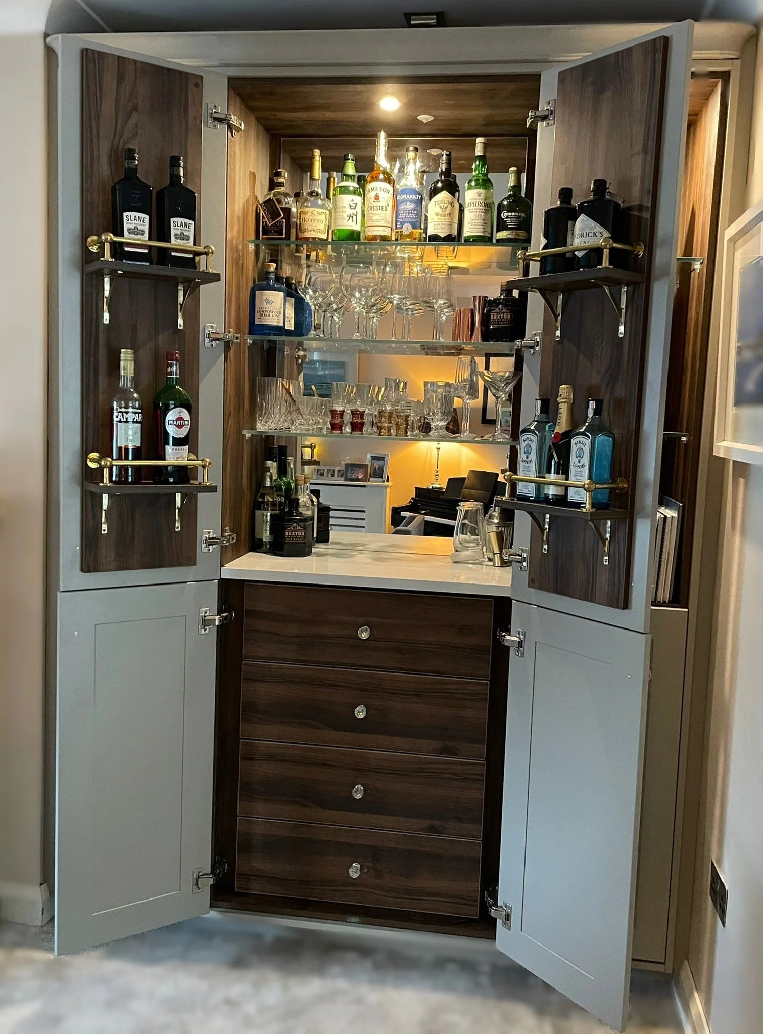 A bar cabinet with open cream-colored doors revealing shelves with various liquor bottles, glassware, and framed photos inside, located in a room with beige walls and carpeted floor.