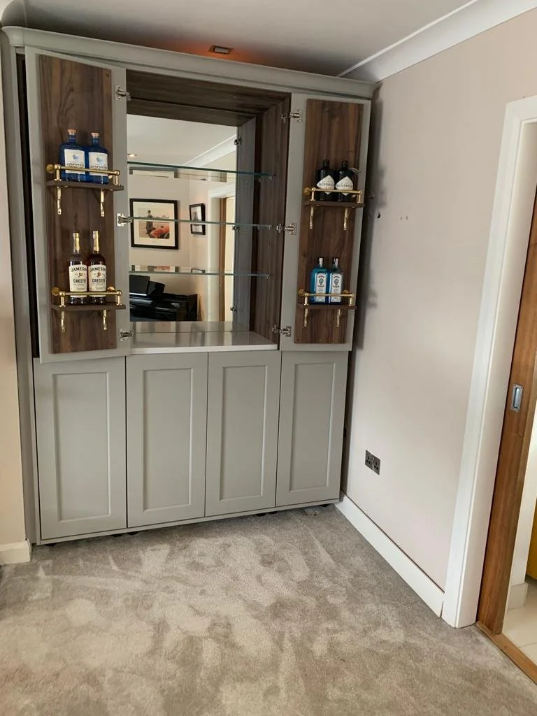 A modern bar cabinet with a mirrored center, glass shelves, and four small brass shelves on either side holding liquor bottles, positioned in a room with beige walls and carpet.