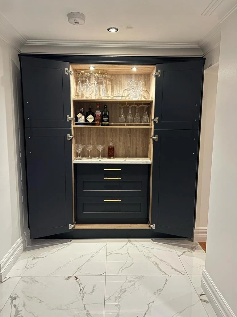 A built-in cabinet with open doors revealing shelves with liquor bottles and glassware, mounted against a wall with white crown molding and marble floor.