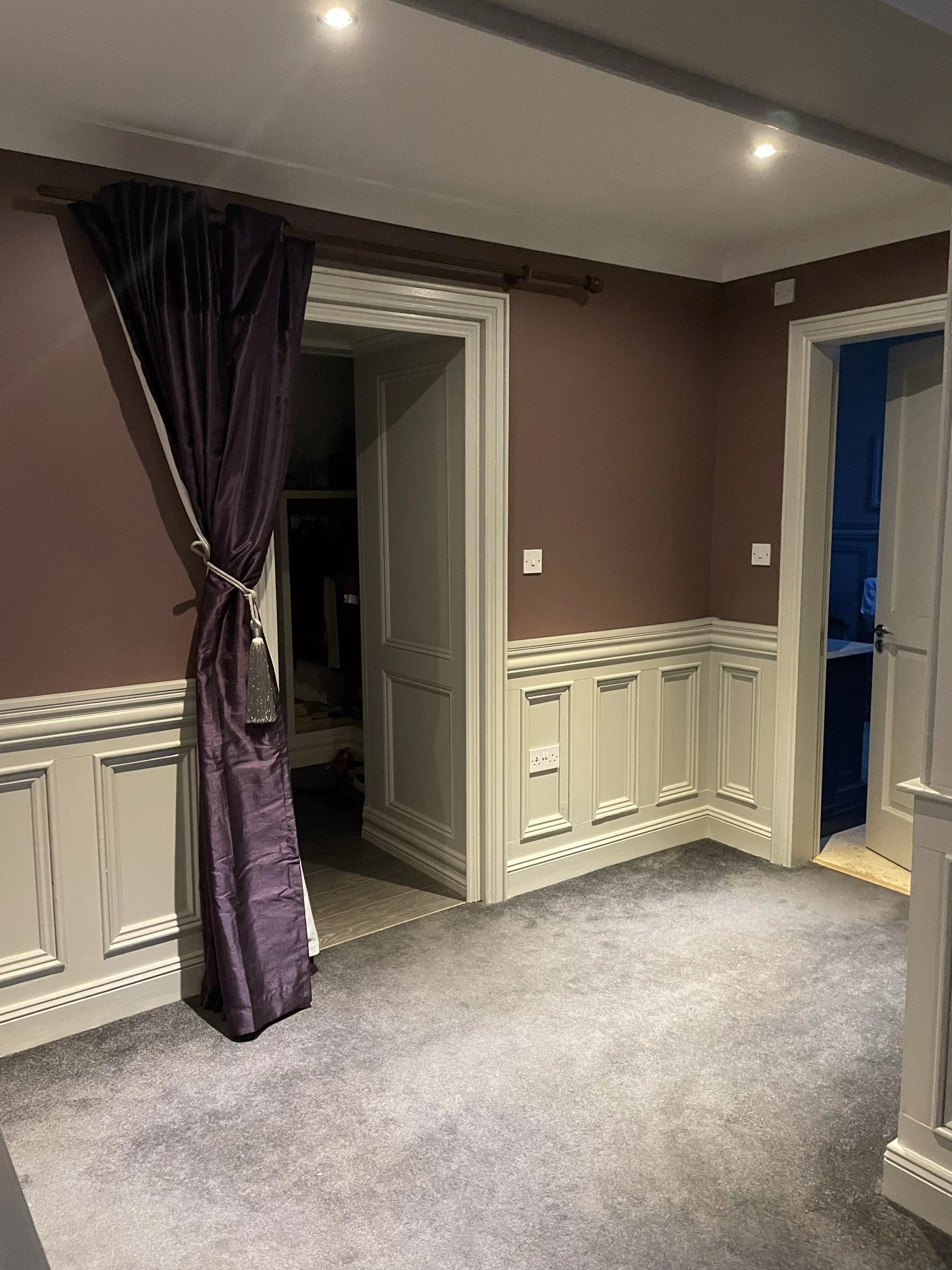 An interior space with beige walls, white wainscoting, gray carpet, and several doorways. A purple curtain is partially pulled to the side, revealing a dark room behind it. Recessed ceiling lights illuminate the area.