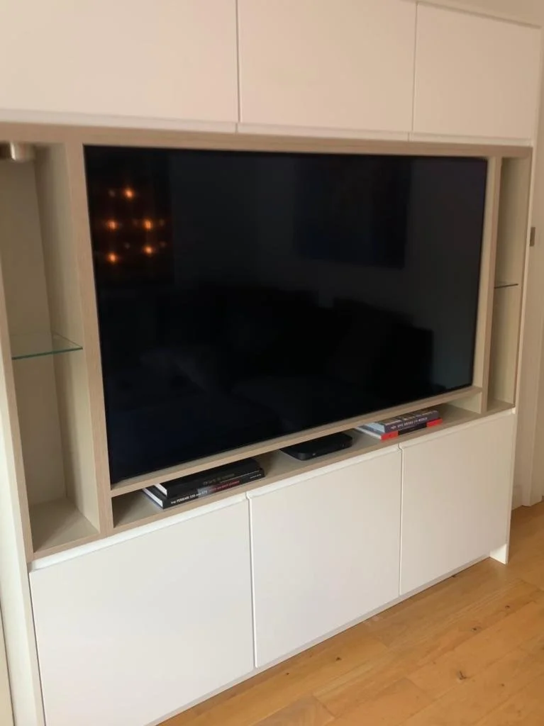 A modern white entertainment center with a flat-screen TV, a glass display shelf on the left, and closed storage below, placed on a wooden floor.