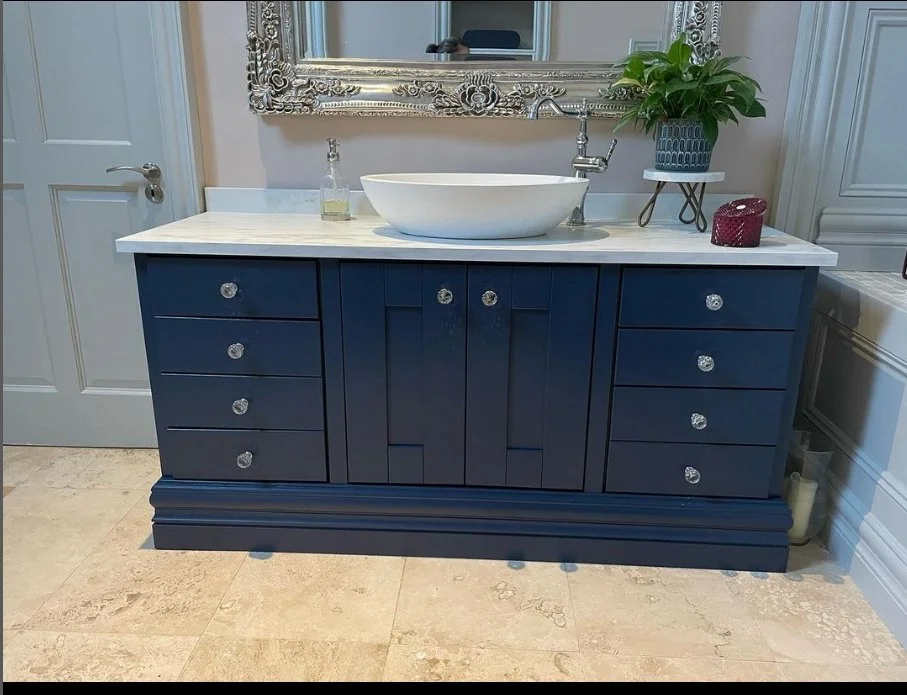 A bathroom vanity with a white countertop, a vessel sink, and a mirror above it. Decor includes a potted plant and small items, set against a styled wall.