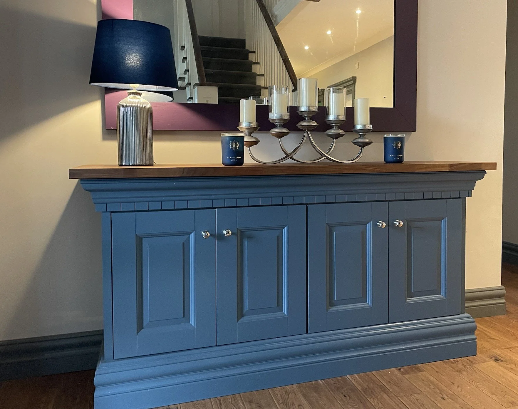 A blue wooden sideboard with decorative molding, topped with a wooden surface. On the surface, there are a black lampshade with a metallic base, a large mirror with a dark wooden frame reflecting part of a staircase, a silver candelabrum with six whi