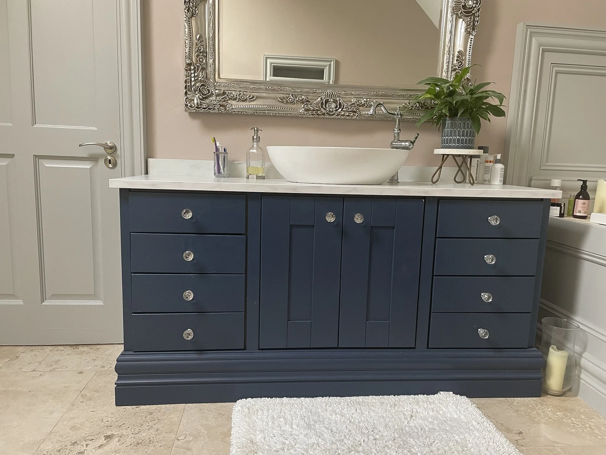 Bathroom with navy blue vanity, white countertop, decorative mirror, plant, soap dispenser, toothbrush holder, and various bottles. Cream-colored tile floor and walls with white door and trim.