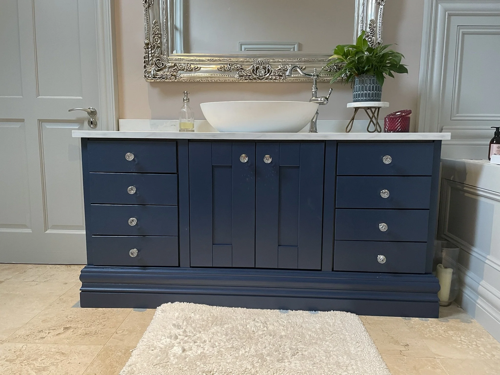A bathroom with a blue vanity featuring multiple drawers with crystal-like knobs, a white countertop with a vessel sink, a yellow soap dispenser, a large decorative mirror with floral detailing, a green potted plant on a small stand, a red container,