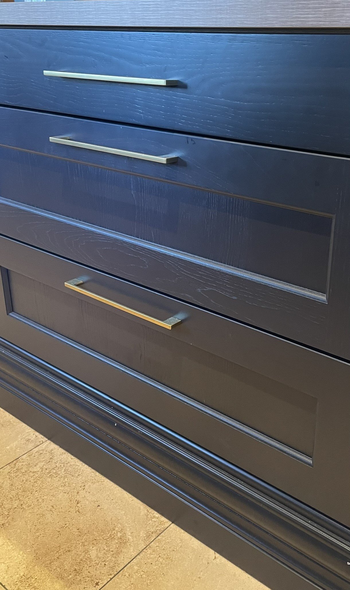 Close-up of a three drawer dresser with silver and gold handles, set on a tiled floor.