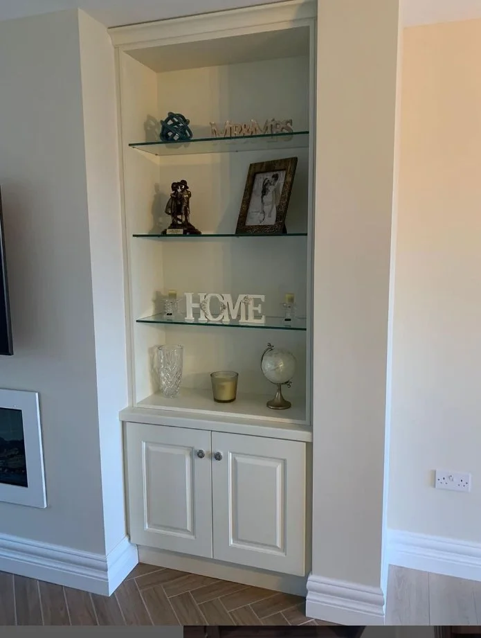Built-in white cabinet with glass shelves displaying decorative items including a framed photo, small sculptures, candles, a glass vase, a candle holder, a globe, and a decorative 'HOME' sign.