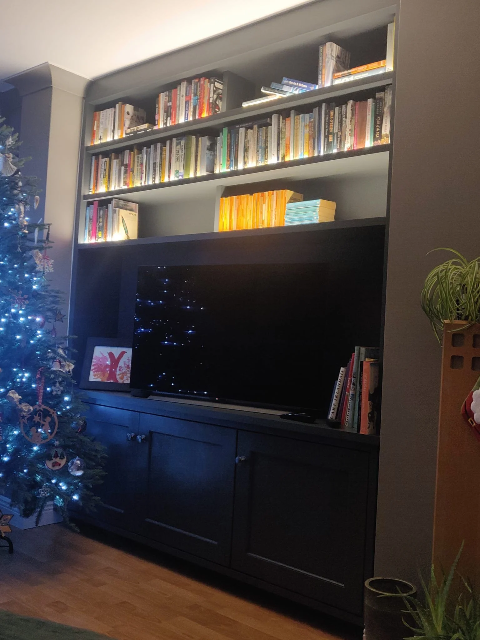A dark gray TV stand with silver knobs, a flat-screen TV on top, a bookshelf with books on the top shelf, and a Christmas tree with blue and white lights and various ornaments on the left side of the image. There is a potted plant in the bottom right