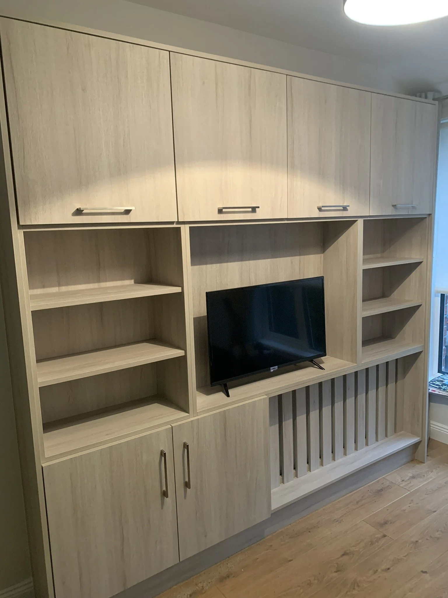 A modern wooden entertainment center with closed cabinets, open shelves, and a small TV, situated on a wooden floor near a window.