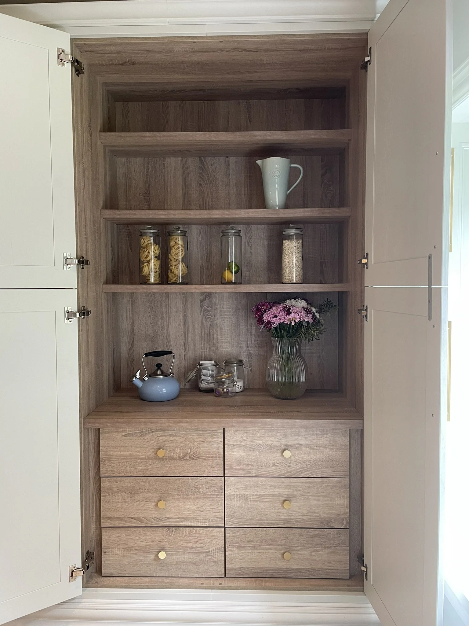 A wooden kitchen cabinet with open white doors revealing shelves and drawers. On the shelves, there is a white pitcher, three glass jars containing pasta, a lime, a lemon, and rice, and a large glass vase with pink and purple flowers on the bottom sh