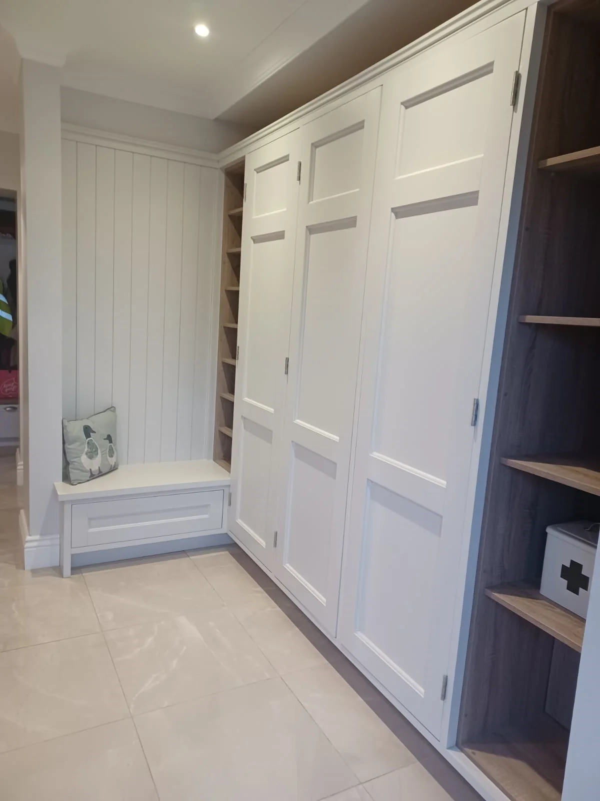 A white built-in storage cabinet and shelving unit in a room with tiled flooring, a cushioned seat with a decorative pillow, and a white medical box on a wooden shelf.