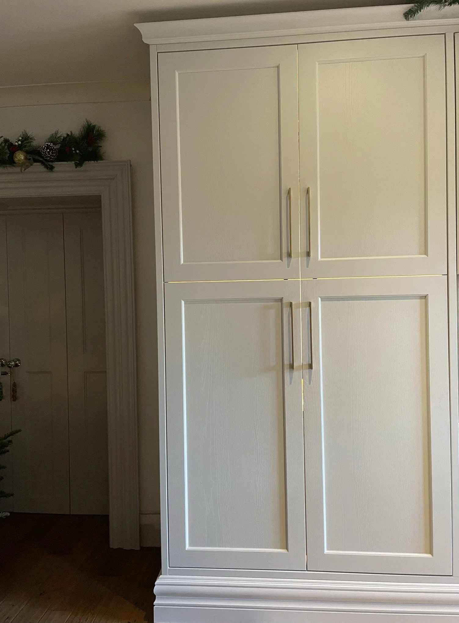 A white cabinet with two doors and gold handles in a room decorated for Christmas with a green garland and ornaments