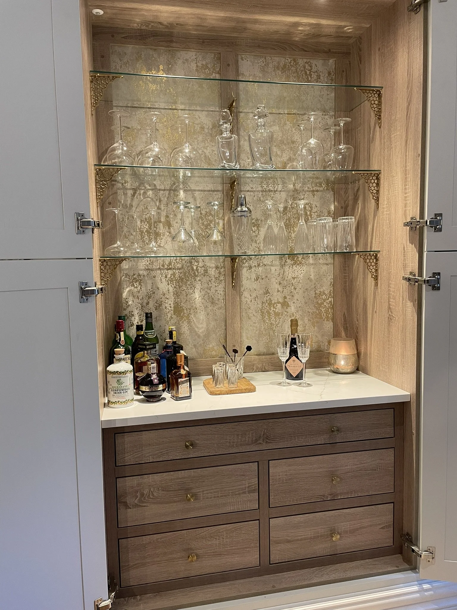 A wooden cabinet with glass shelves inside, displaying wine glasses and liquor bottles, with a bar setup on the countertop below.