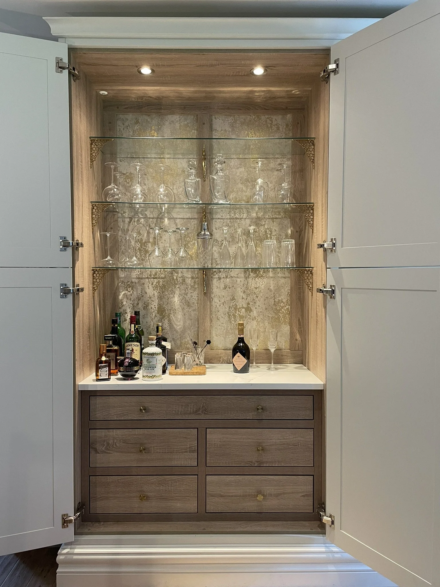 Open cabinet with glassware and liquor bottles, bar accessories, and glasses inside, illuminated by ceiling lights.