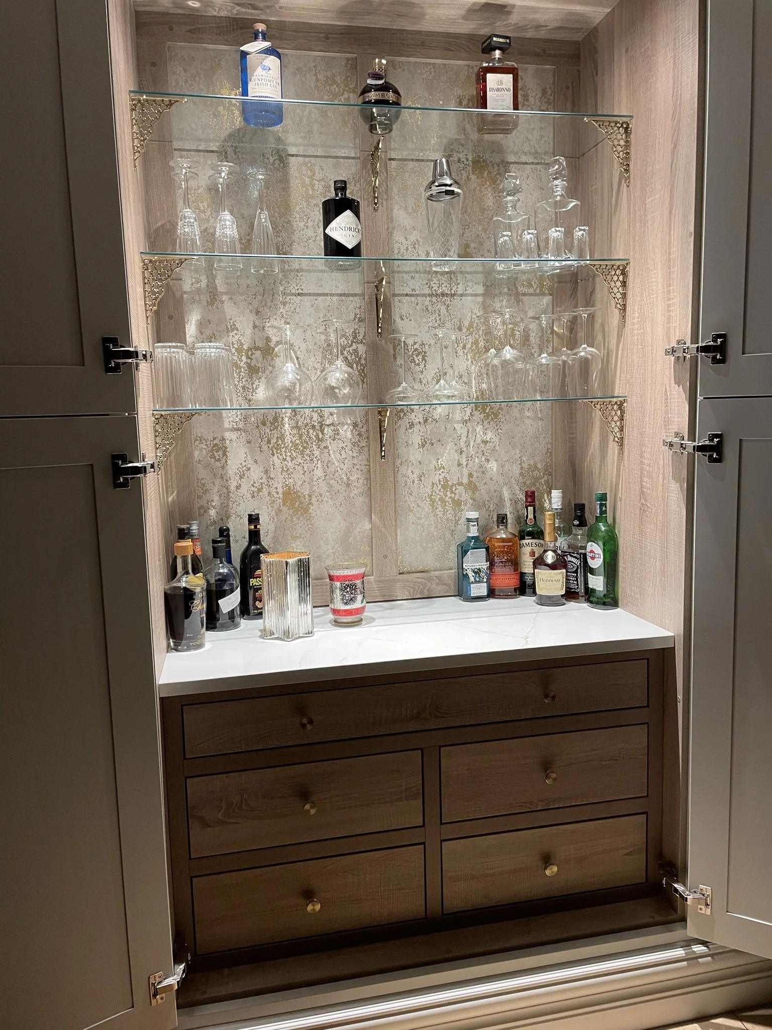 A wooden cabinet with glass shelves inside, containing bottles of alcohol and glassware, and an open door revealing the interior.