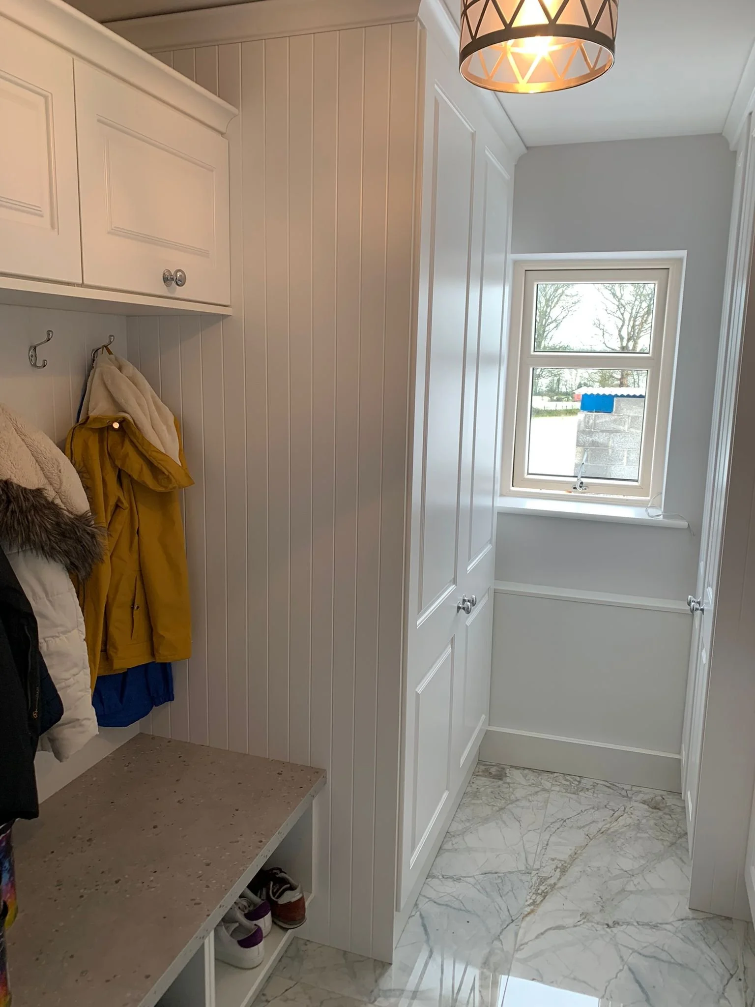 Corner of a white closet with hooks holding jackets and shoes underneath. Bright room with large window, marble floor, and ceiling light.