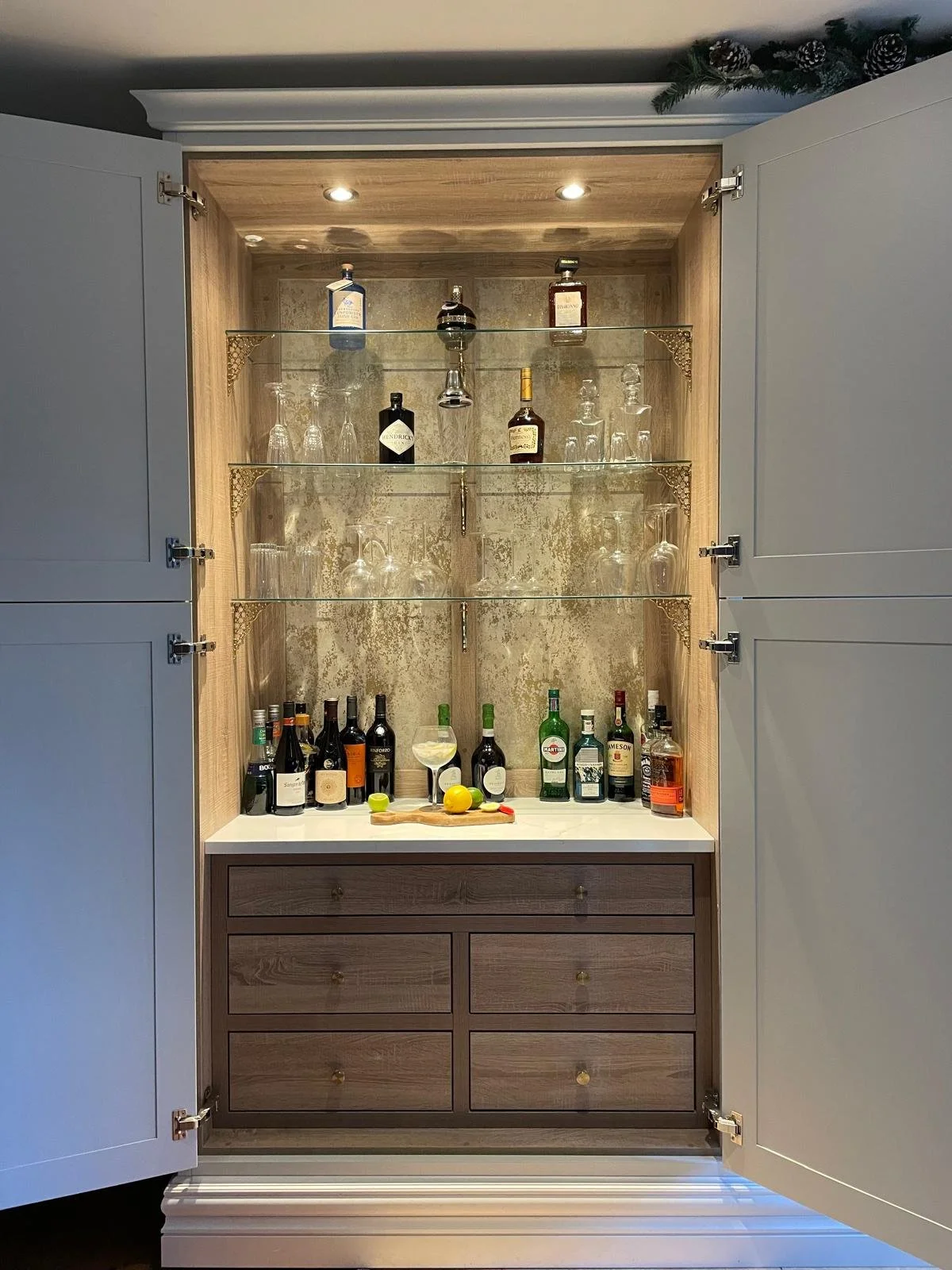 A built-in bar cabinet with glass shelves containing alcohol bottles, glasses, and bar accessories, with a countertop holding more bottles, a glass with a drink, and citrus fruits. The cabinet has closed white doors on the sides and a decorative gold-ornamented glass back panel.