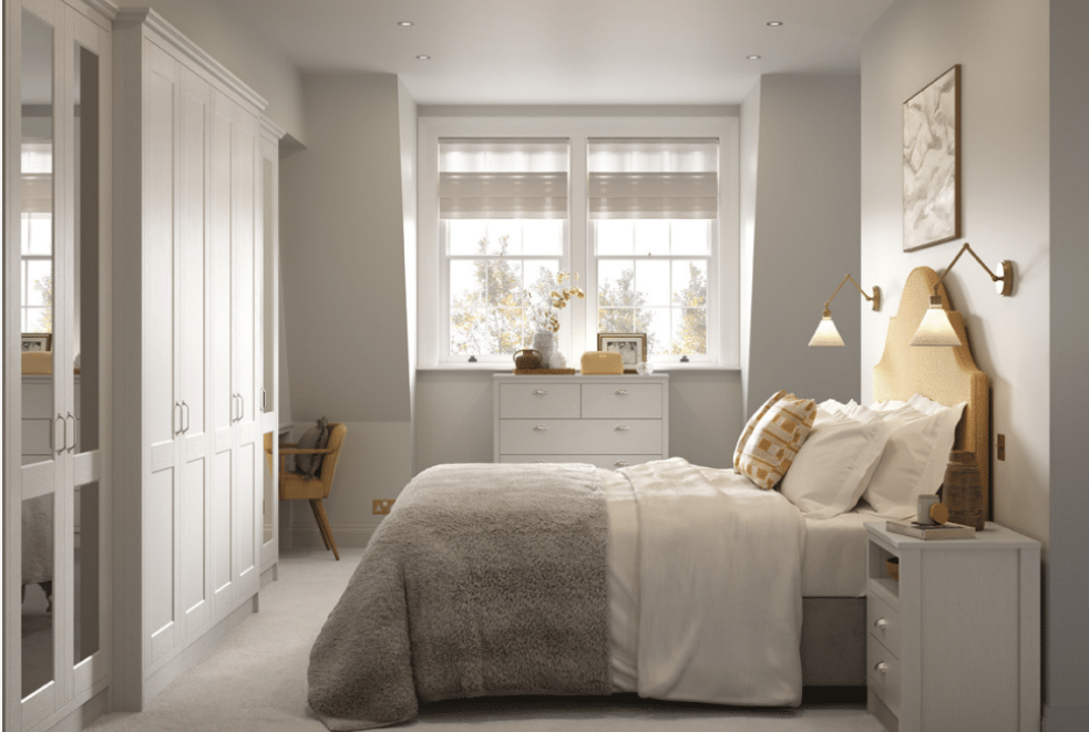 Bright bedroom with white walls, a large bed with white bedding and a beige headboard, modern wall-mounted lamps, a white dresser below windows, and a closet with mirrored doors.