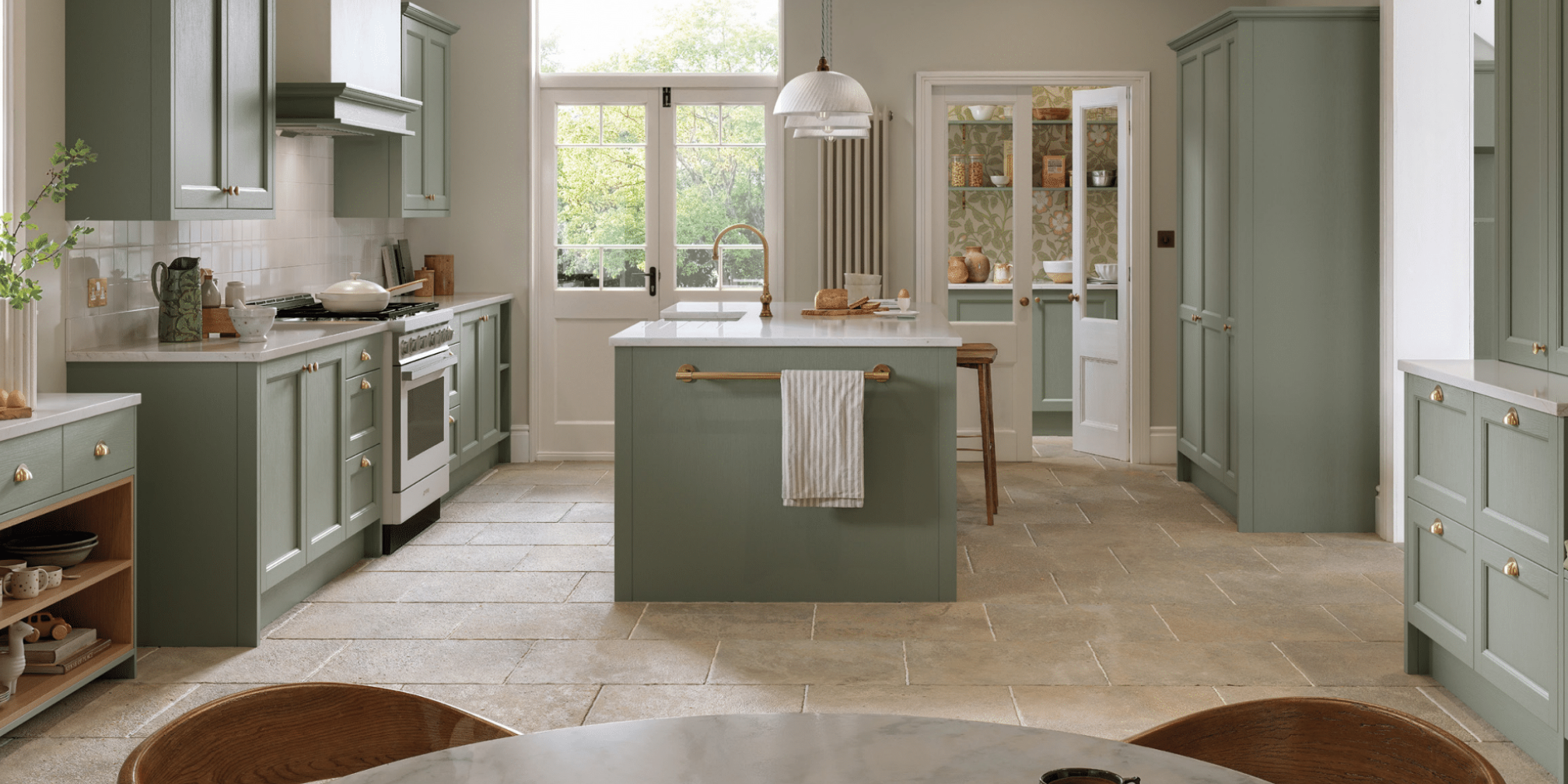 A spacious kitchen with green cabinetry, a central island, and large windows overlooking trees. The kitchen features a white countertop, a sink with a gold faucet, and a dining area with wooden chairs.