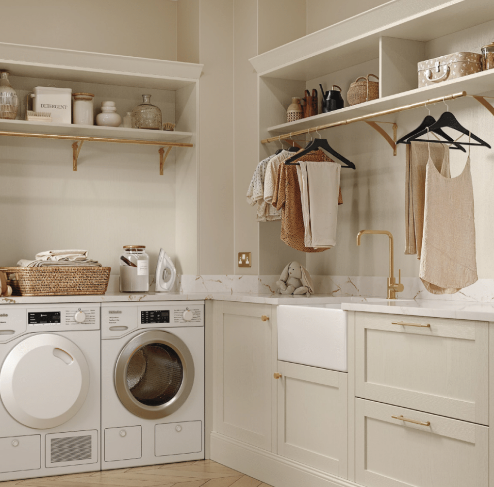 Laundry room with white washer and dryer, open shelves with storage jars, woven basket, and laundry supplies, beige cabinets with gold handles, a marble countertop, a white farmhouse sink, hanging clothes, and decorative baskets and containers.