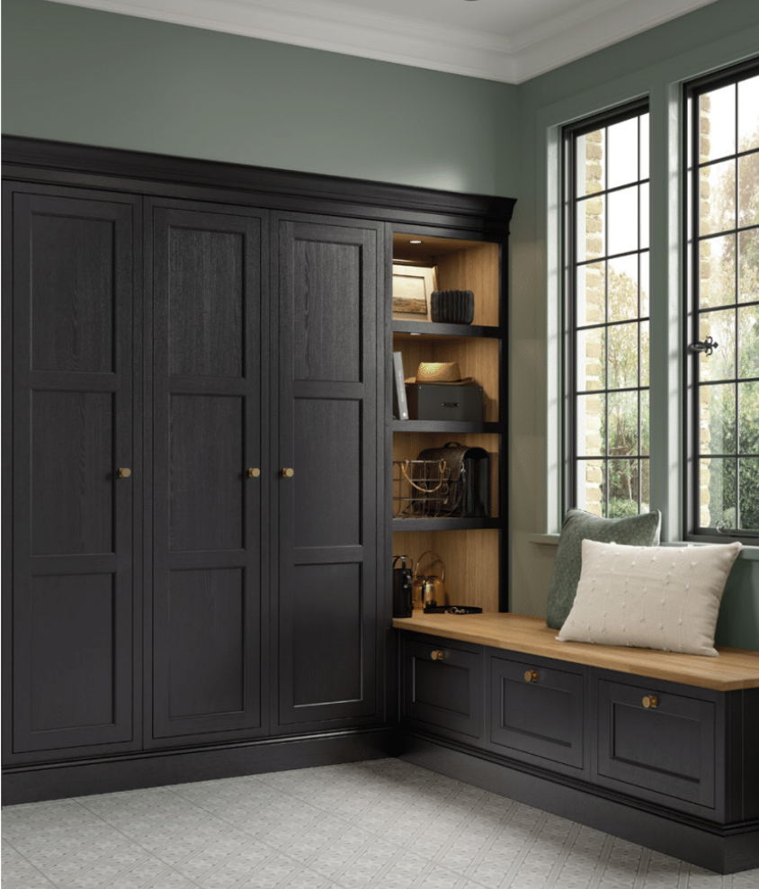 Interior of a room with a large black wooden cabinet, built-in bookshelves, a window seat with pillows, and a window showing trees outside.