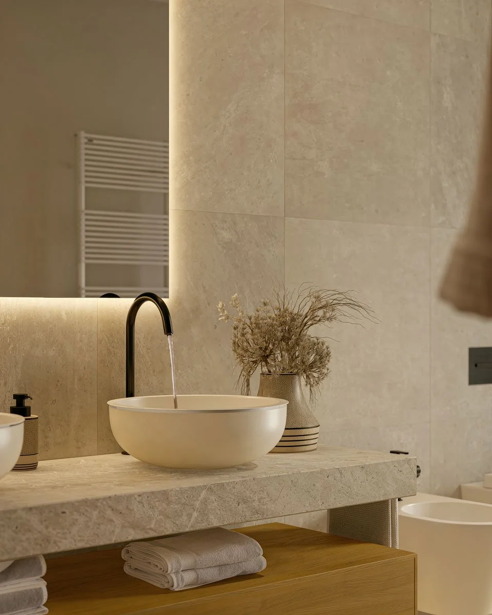 Un bagno moderno con lavabo rotondo bianco, un rubinetto nero e una pianta secca in una lustra. Sul piano ci sono asciugamani bianchi piegati e un dispenser. Sullo sfondo si scorge uno specchio e una parete rivestita in pietra beige.