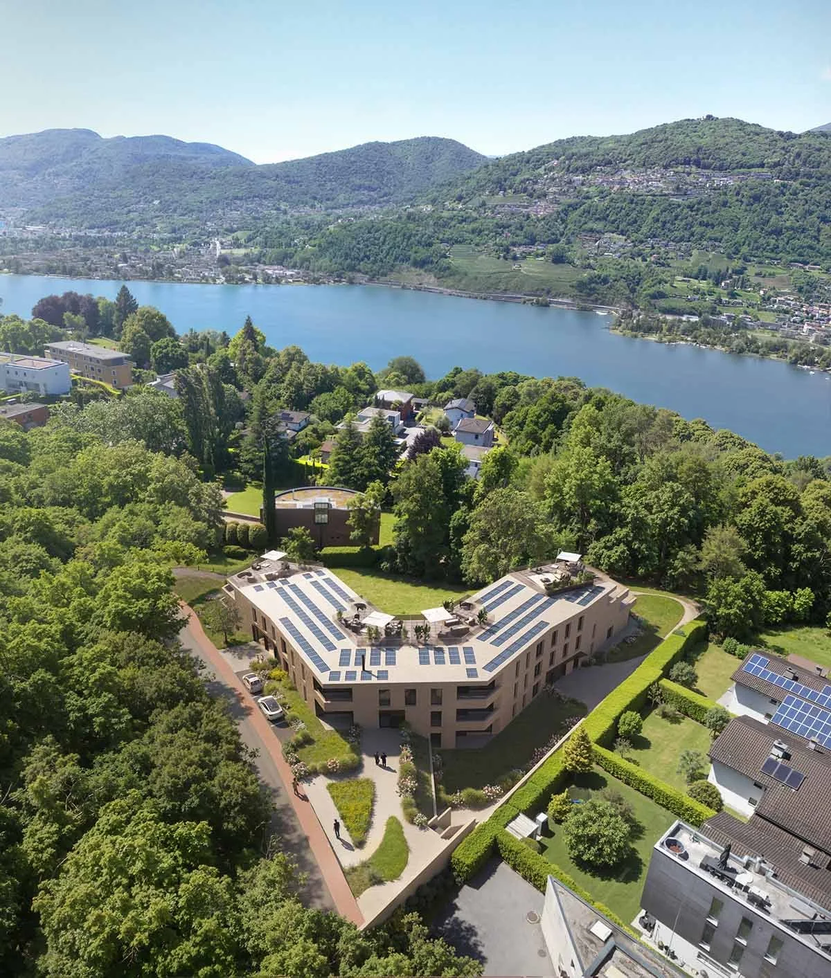 Vista aerea di un edificio residenziale con pannelli solari sul tetto, circondato da alberi e con un lago e montagne sullo sfondo.