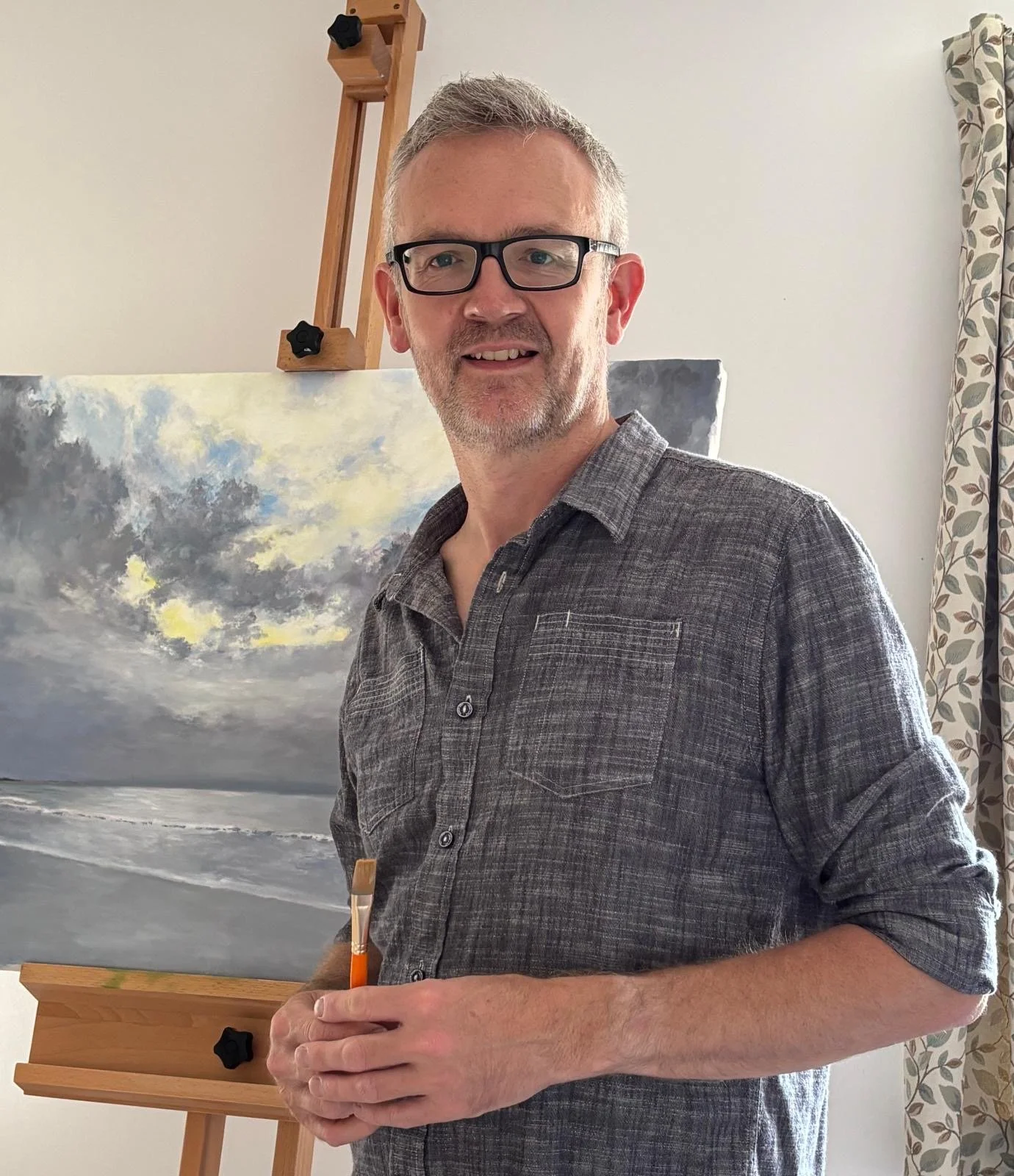 A man with gray hair, glasses, and a gray plaid shirt standing next to an easel with a painting of a cloudy sky over the ocean.