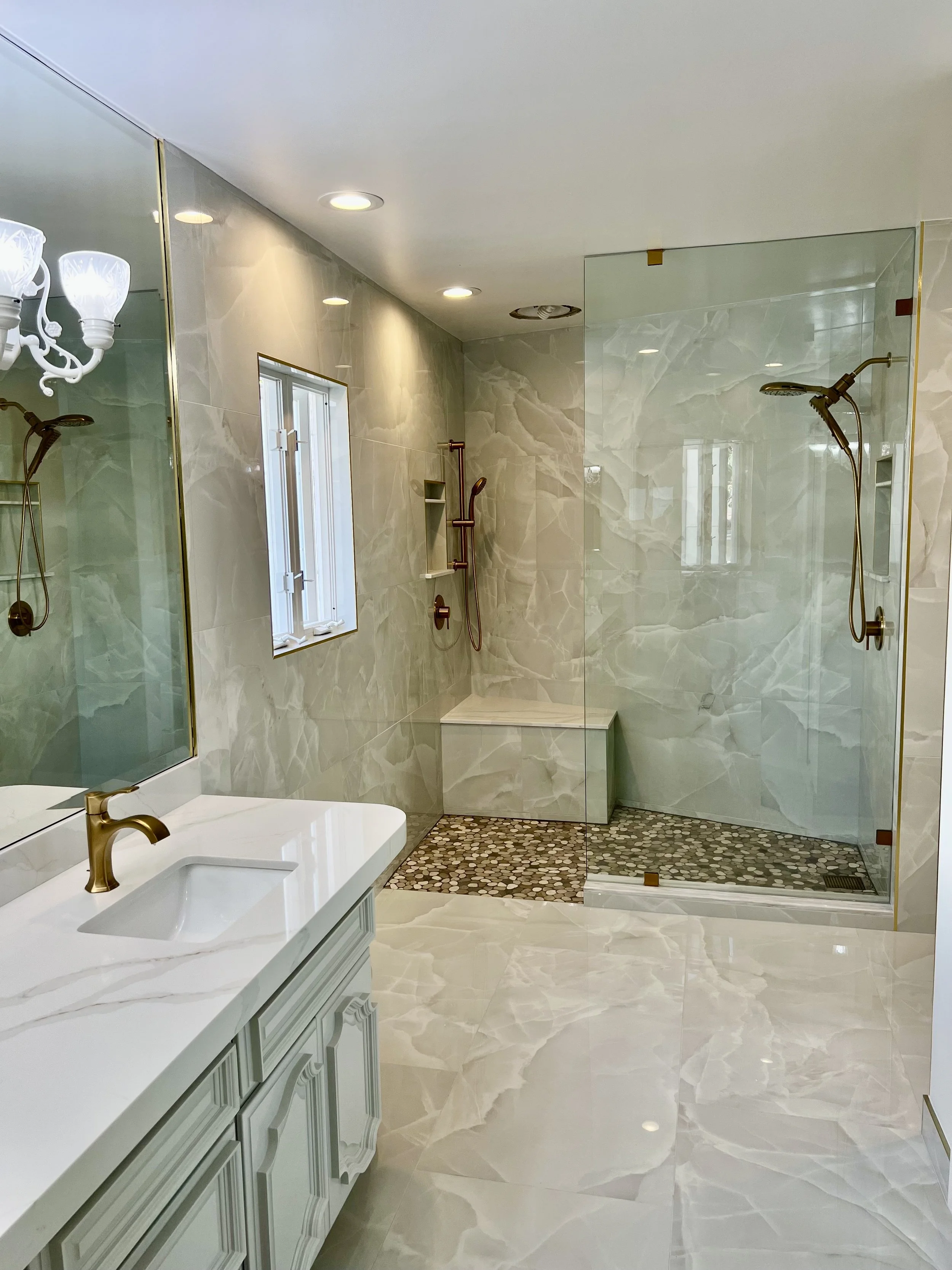 “This walk-in shower features floor-to-ceiling tile, a level entry with pebble flooring that is flush with the rest of the bathroom floor, a rain shower on the left, a bench, two soap niches, and a fixed glass panel above the curved enclosure.”
