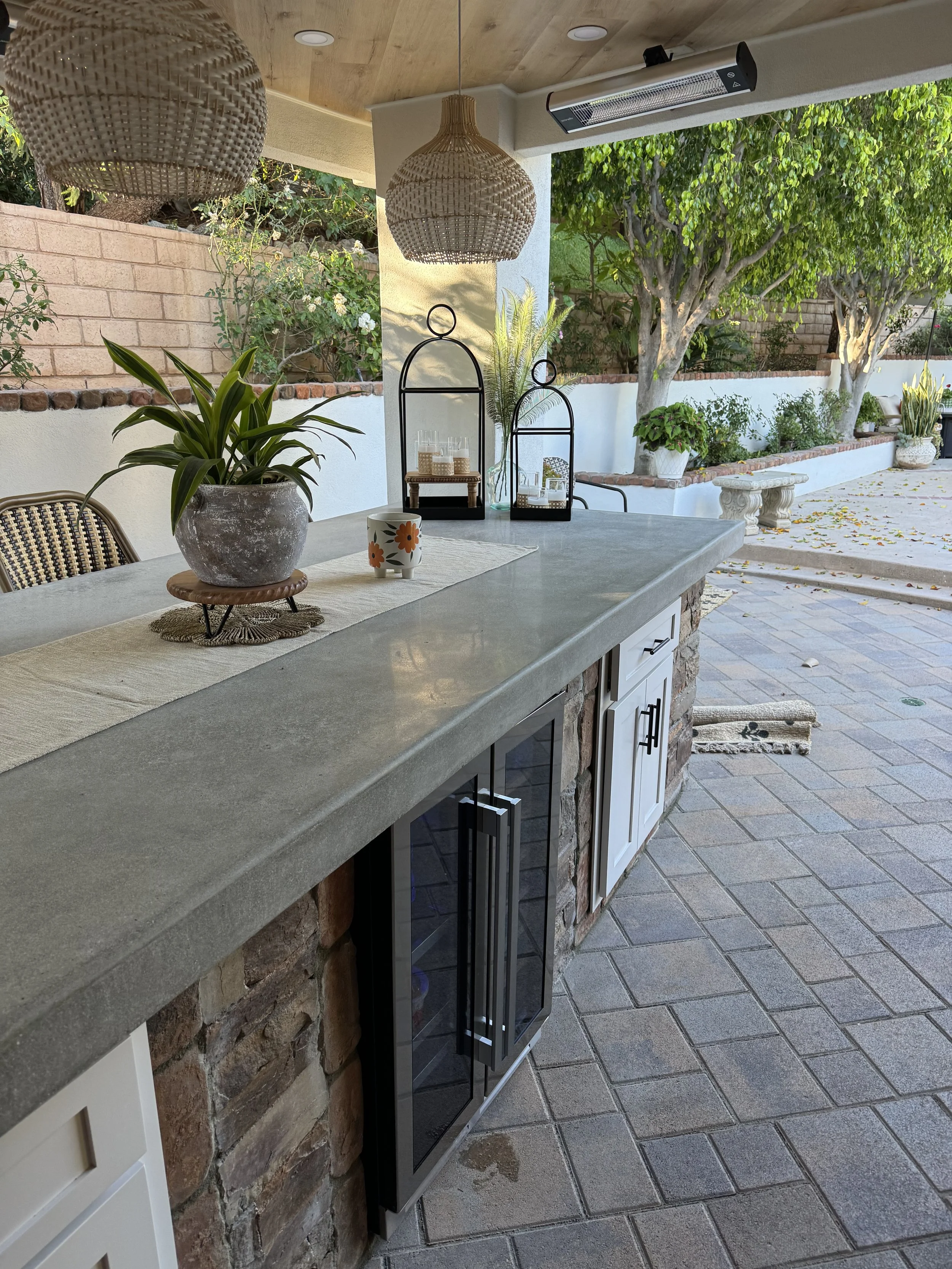 “This custom-built exterior bar features a concrete countertop, built from scratch, with a wine refrigerator installed, side power outlets, and two inset cabinets. The bar is accented with stacked stone, creating a sleek, modern outdoor entertaining