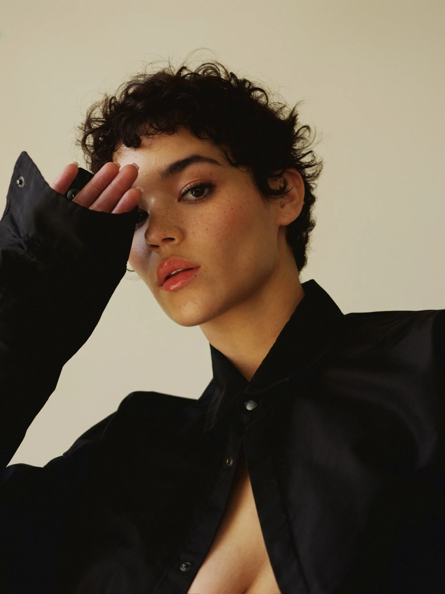 A woman with short, curly dark hair and freckles, wearing a black shirt, covering part of her face with her left hand, which has long fingers and a black sleeve, against a plain beige background.