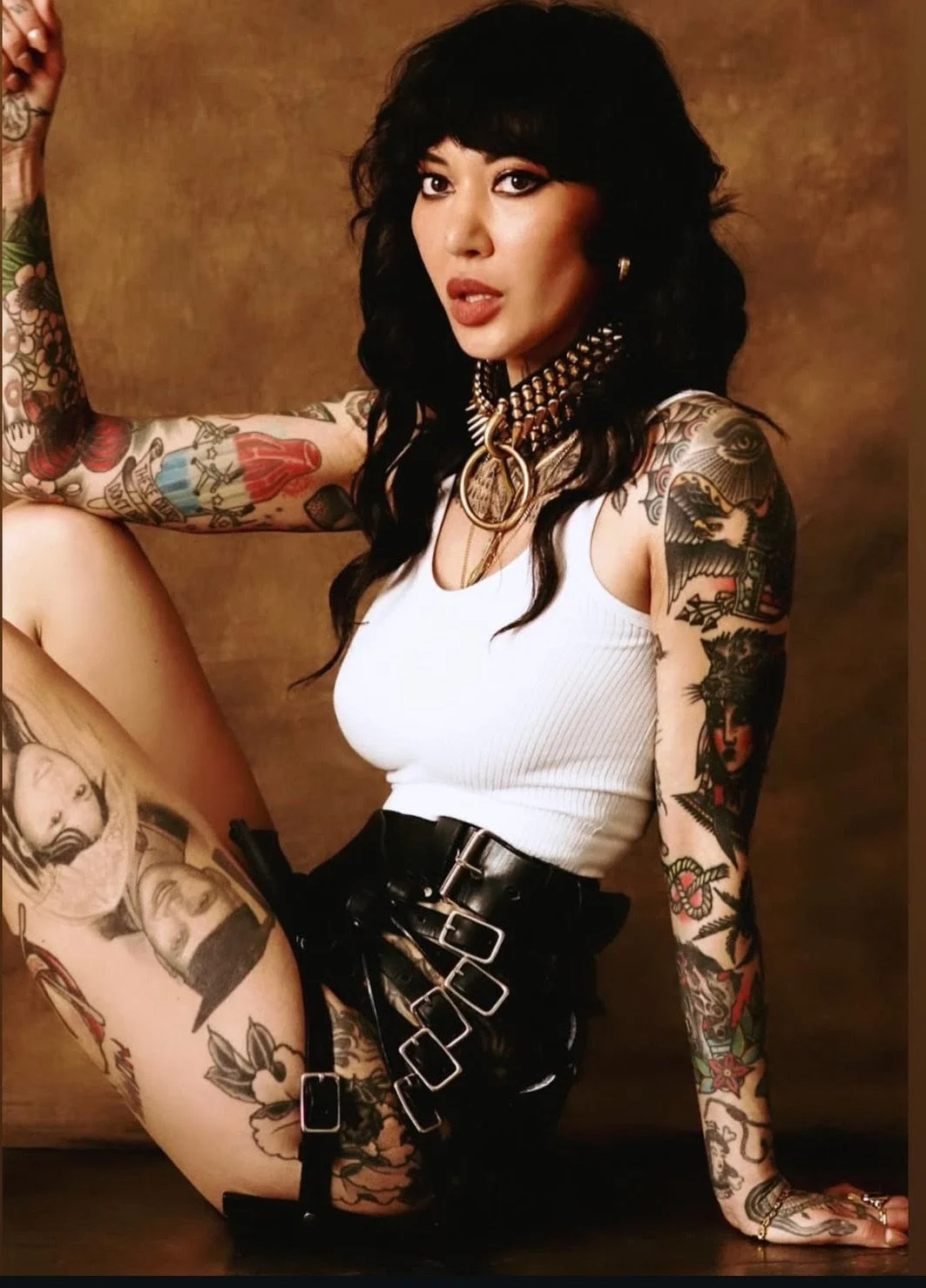 A woman with black hair and tattoos sitting against a brown background, wearing a white top and black belt shorts with multiple buckle straps.
