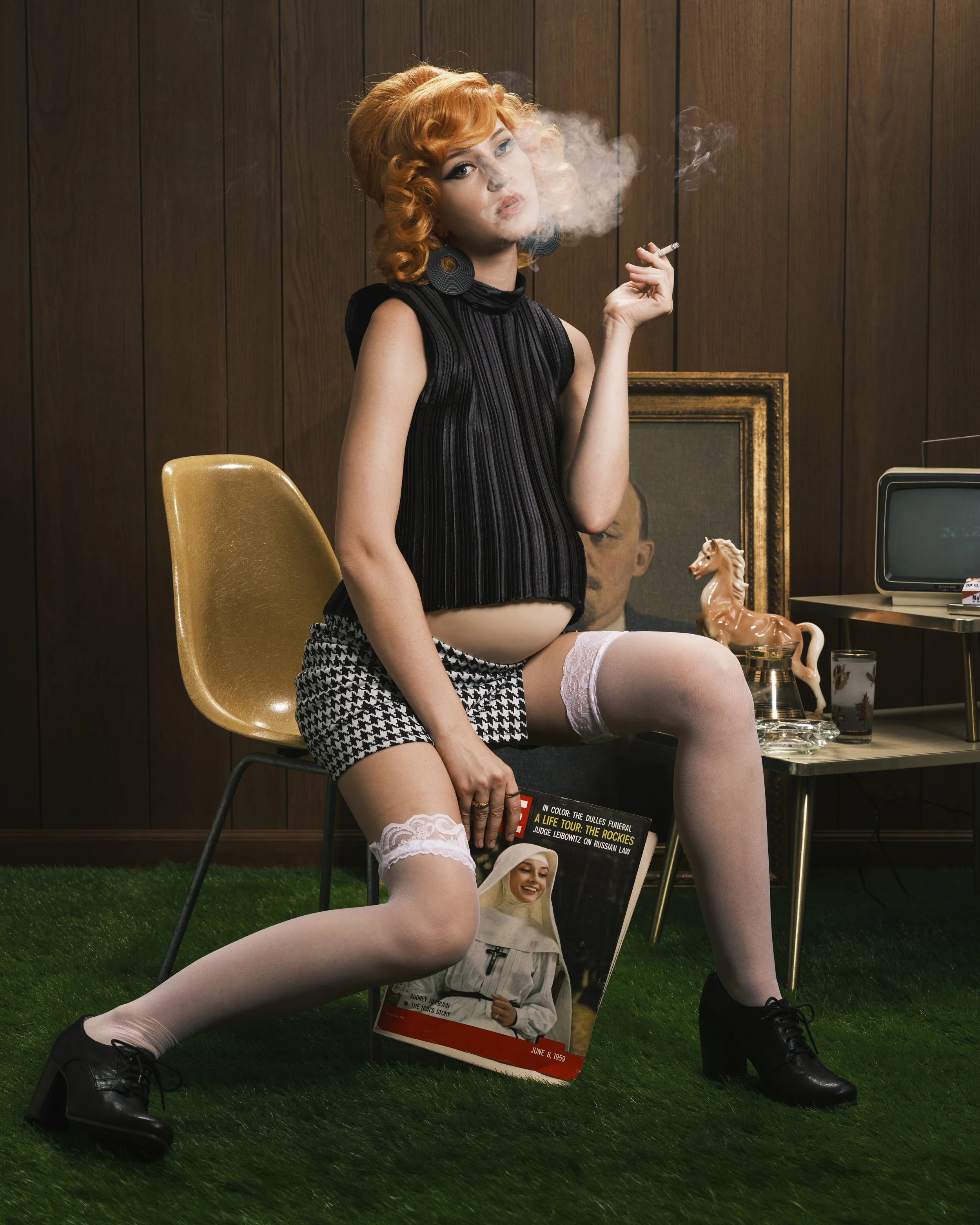 A woman with red curly hair and vintage style makeup sitting on a chair in front of a wood-paneled wall, smoking a cigarette, wearing a black vintage sleeveless top, houndstooth shorts, white lace thigh-high stockings, and black shoes. She holds a ma