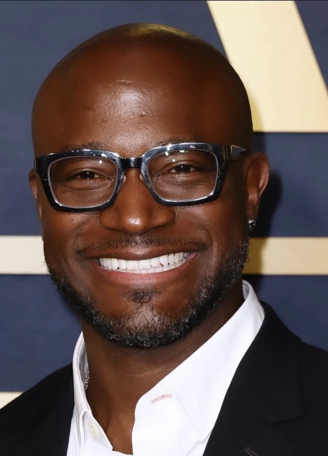 A smiling man wearing glasses, a white shirt, and a dark blazer, posed in front of a dark background with cream-colored stripes.